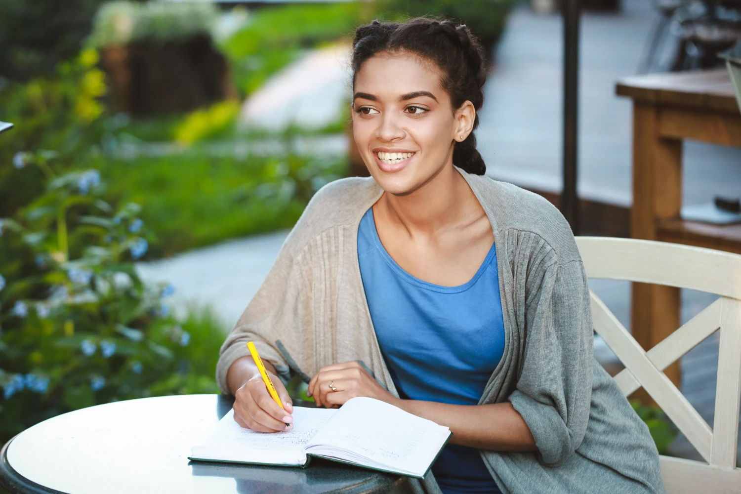 Jovem mulher sorridente escrevendo em um caderno enquanto está sentada em uma mesa ao ar livre, em um ambiente verde.