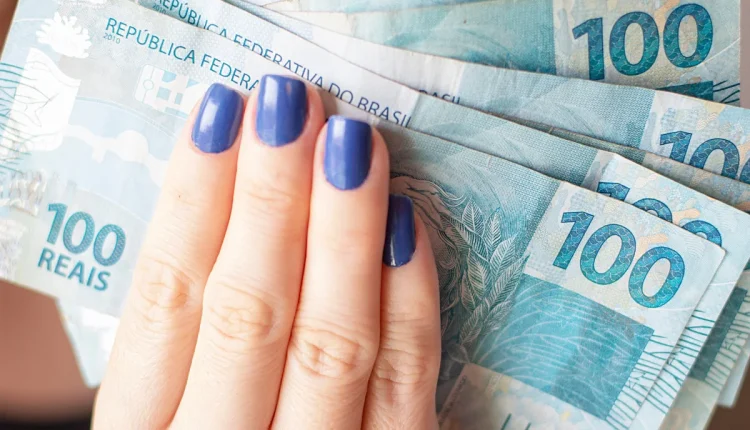 mão feminina com unhas pintadas segurando várias notas de 100 reais.