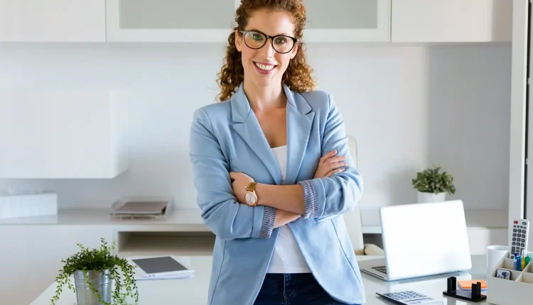 Mulher sorridente com óculos e blazer azul claro, transmitindo confiança em ambiente de escritório moderno.