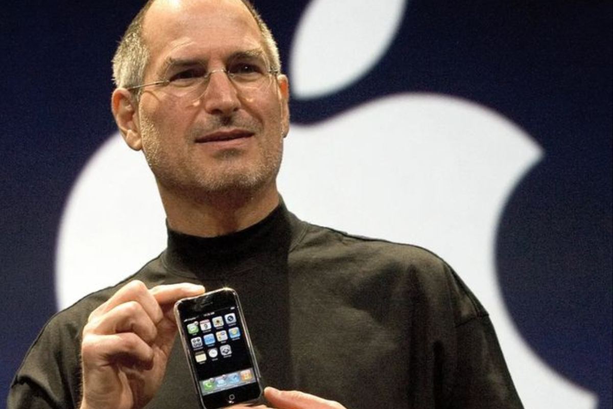 Steve Jobs segurando um smartphone com o logotipo da Apple ao fundo.