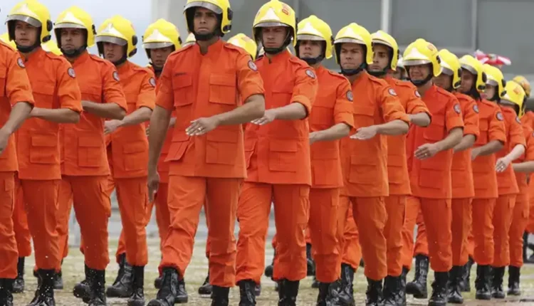Grupo de bombeiros marchando durante treinamento com uniforme completo, incluindo capacetes amarelos e trajes de combate à incêndio.