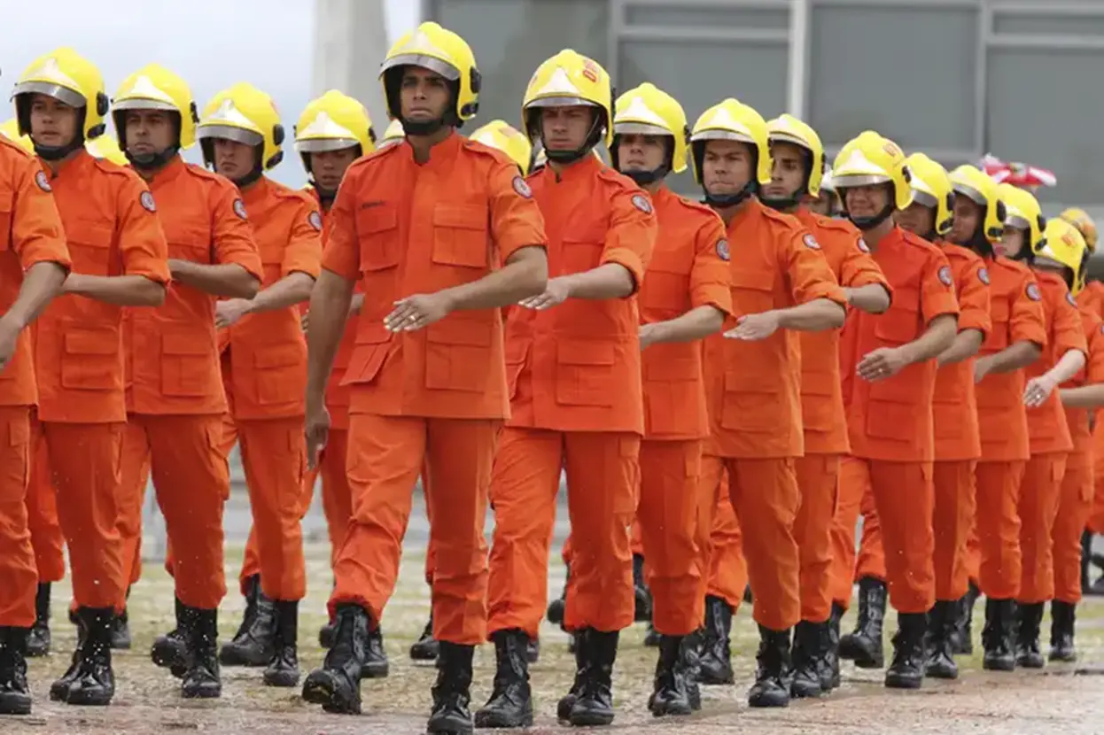Grupo de bombeiros marchando durante treinamento com uniforme completo, incluindo capacetes amarelos e trajes de combate à incêndio.