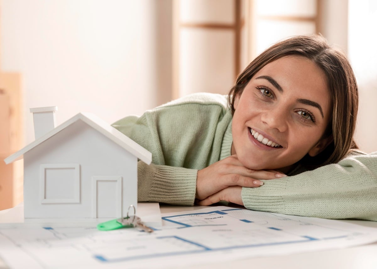 Mulher sorridente ao lado de uma maquete de casa e chave sobre planta baixa, representando realização de sonho com financiamento habitacional.