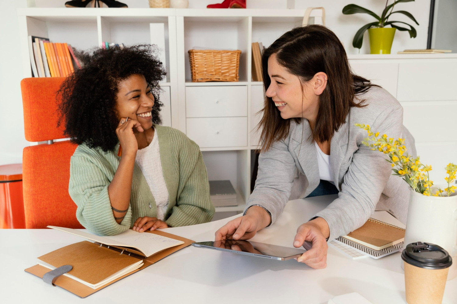 Duas pessoas sorrindo e compartilhando ideias, uma com um tablet e outra com um caderno, em um ambiente de trabalho criativo.