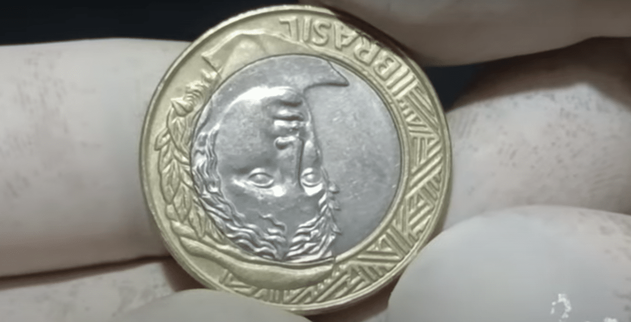 Moeda de 1 real com reverso invertido sendo segurada por luvas