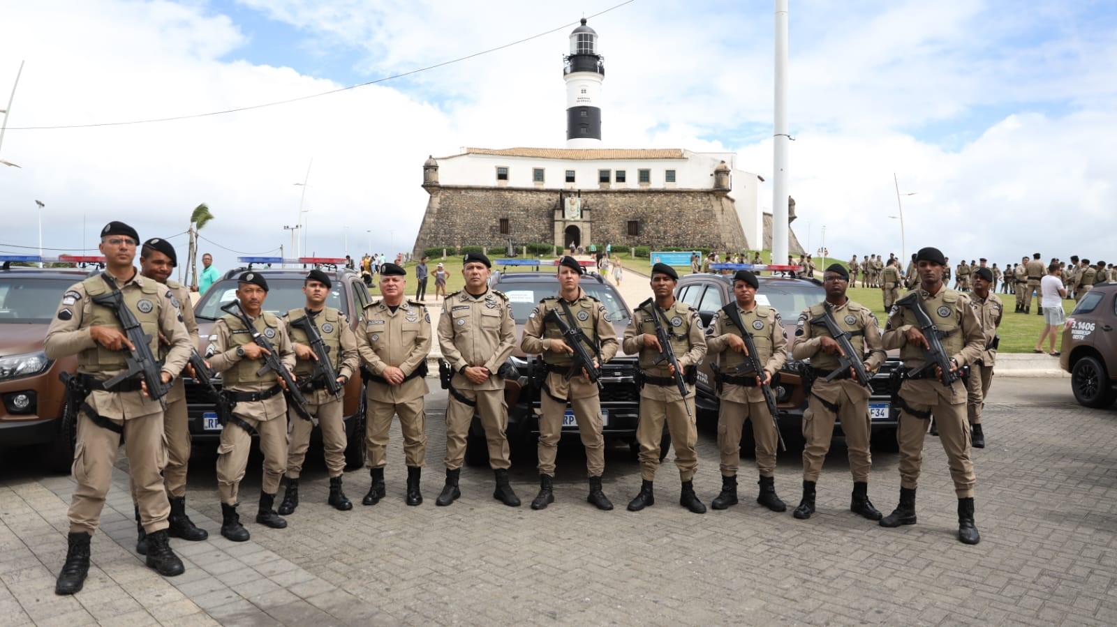 Policiais militares posando com veículos e o Farol da Barra ao fundo em Salvador.