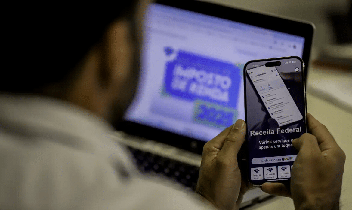 Pessoa acessando o site da Receita Federal pelo celular com o Imposto de Renda aberto no computador.