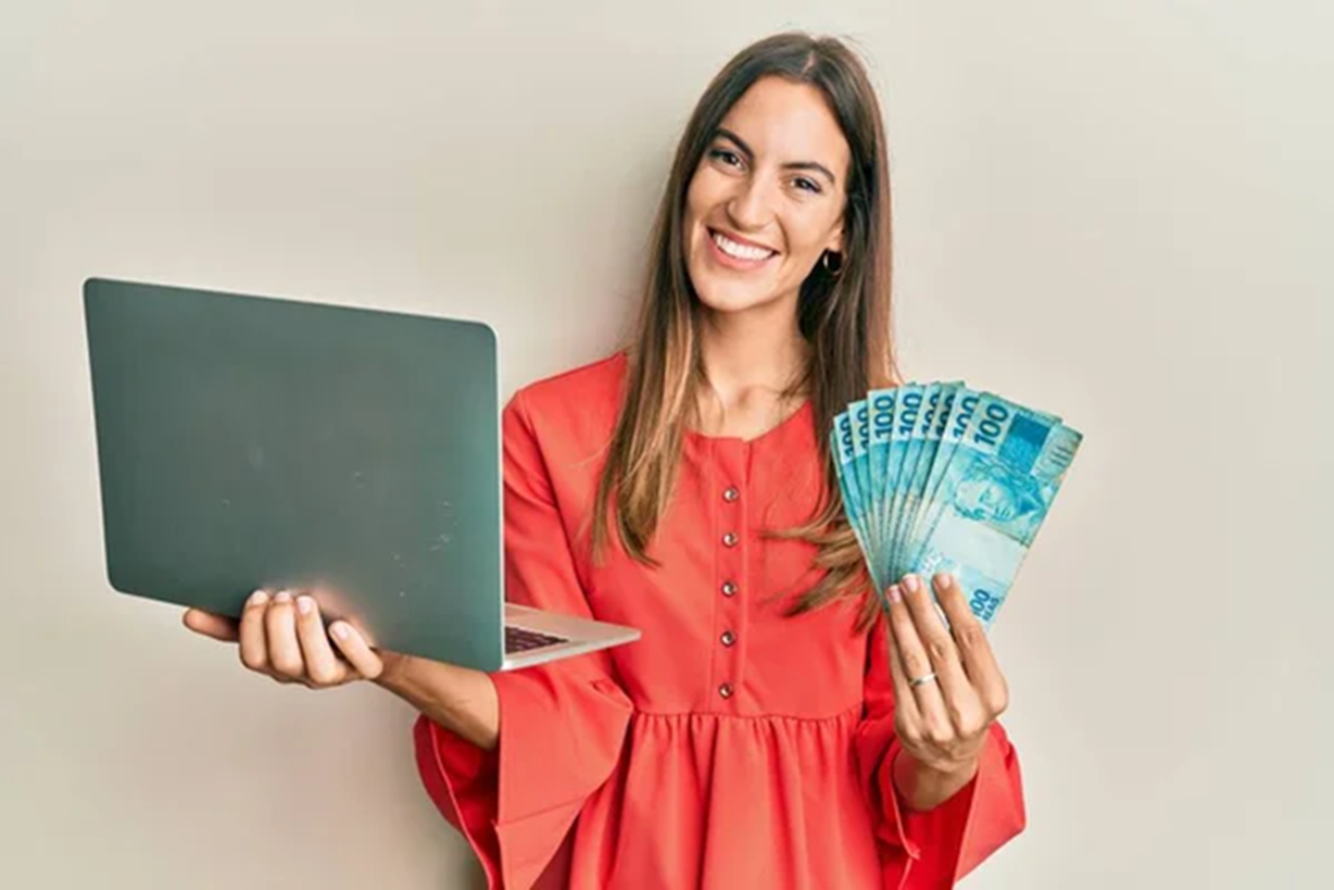 Mulher segurando dinheiro e um laptop, simbolizando o saque de até um salário mínimo e o processo de recebimento de benefícios.