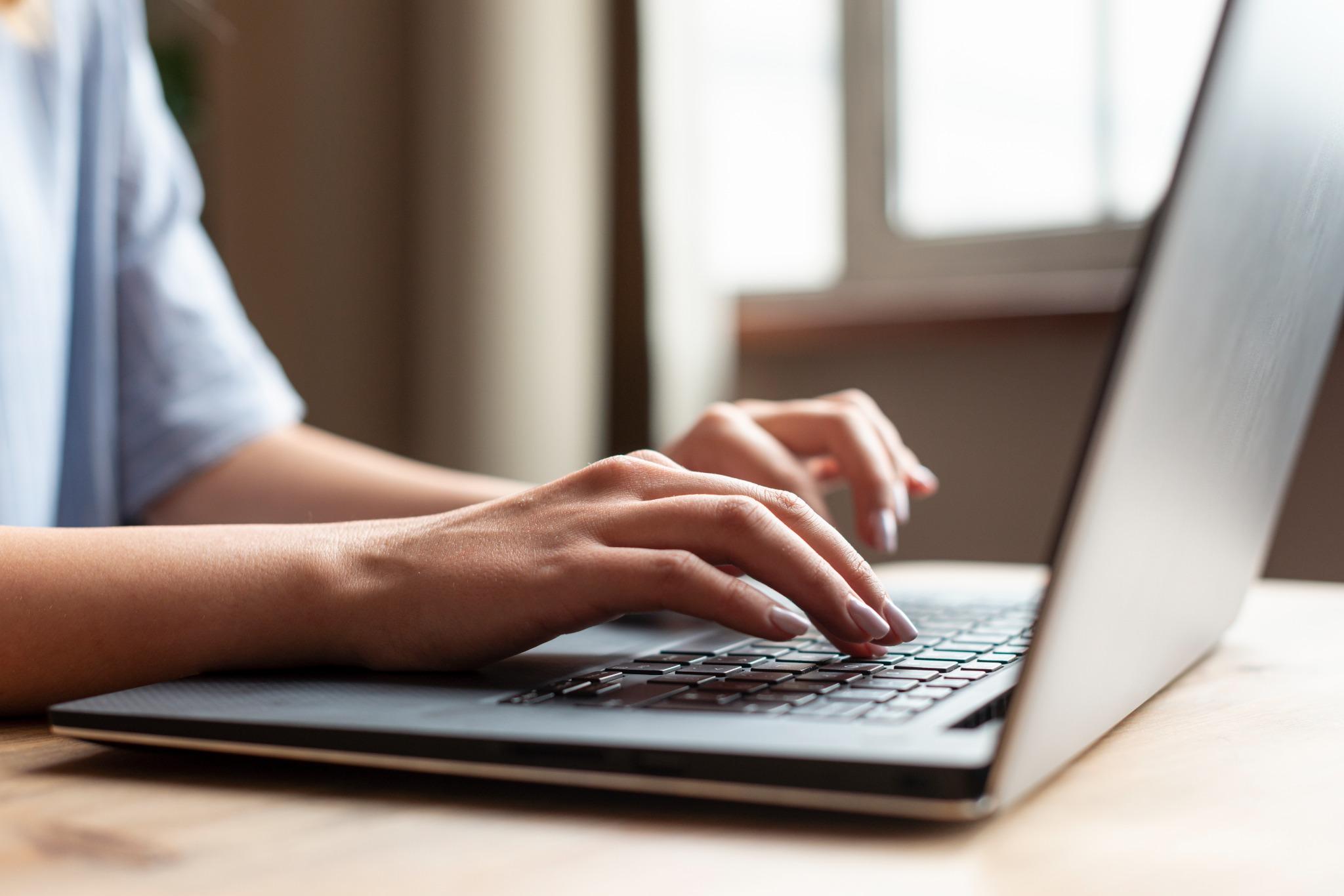 Mãos femininas digitando em teclado de notebook sobre mesa de madeira.