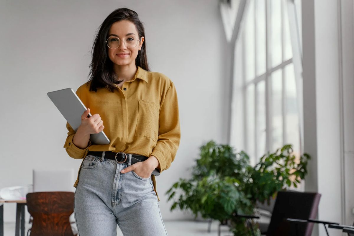 Jovem empresária sorrindo e posando com um tablet em um ambiente de trabalho moderno e iluminado.