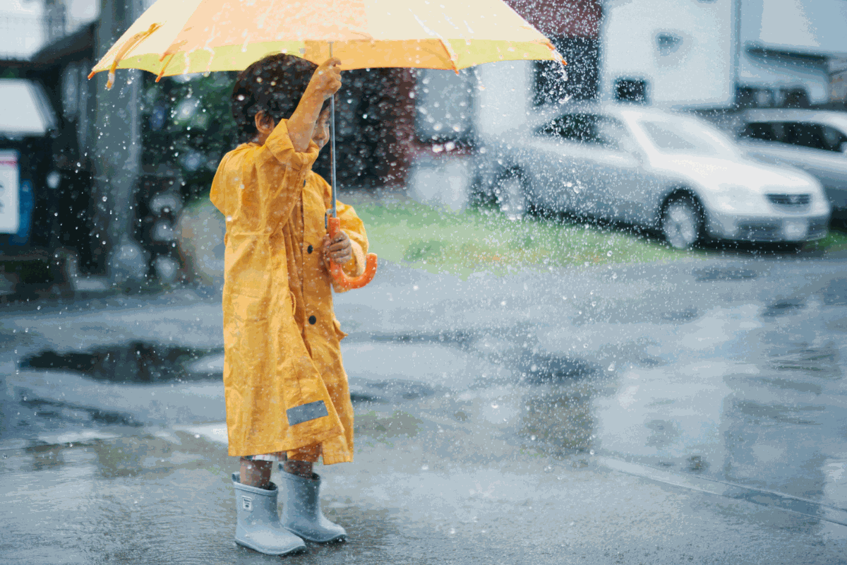 Criança com capa de chuva amarela segurando guarda-chuva colorido em dia chuvoso.
