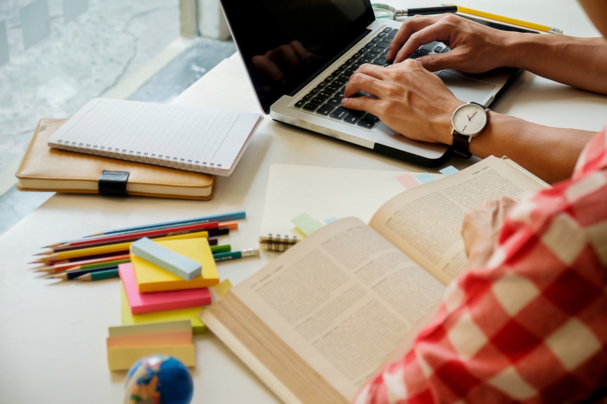 Pessoa estudando com laptop, livro aberto e materiais escolares sobre a mesa.