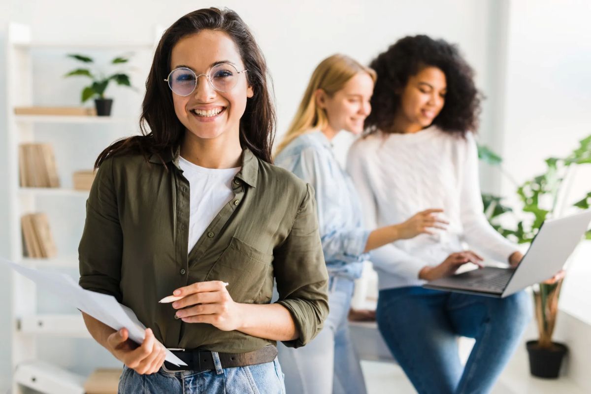 Grupo de mulheres colaborando no ambiente de trabalho, com uma delas sorrindo enquanto segura papéis e as outras usando um laptop.