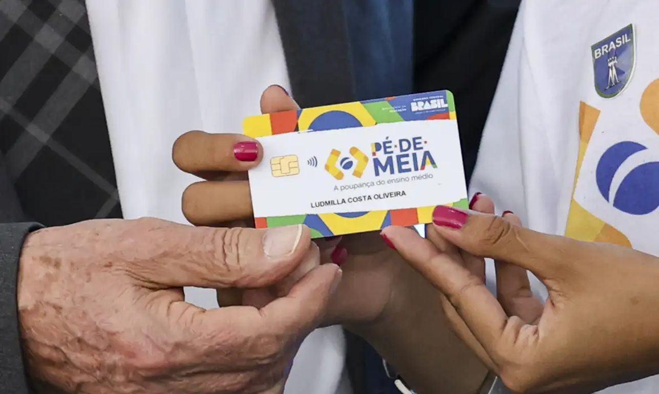 Cartão do programa Pé-de-Meia sendo mostrado por duas pessoas, simbolizando o pagamento e benefício do programa.