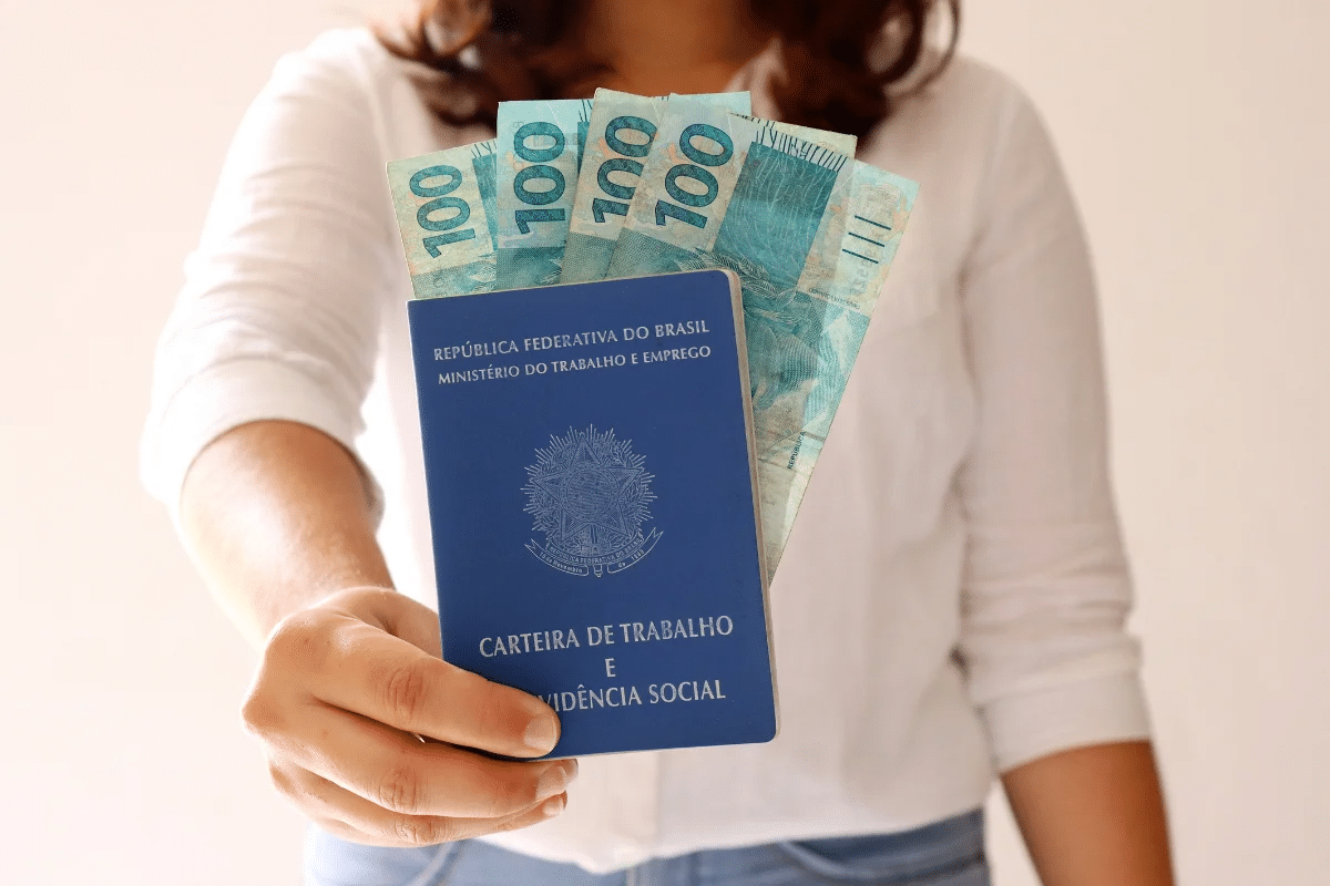 Mulher segurando carteira de trabalho com notas de R$ 100.