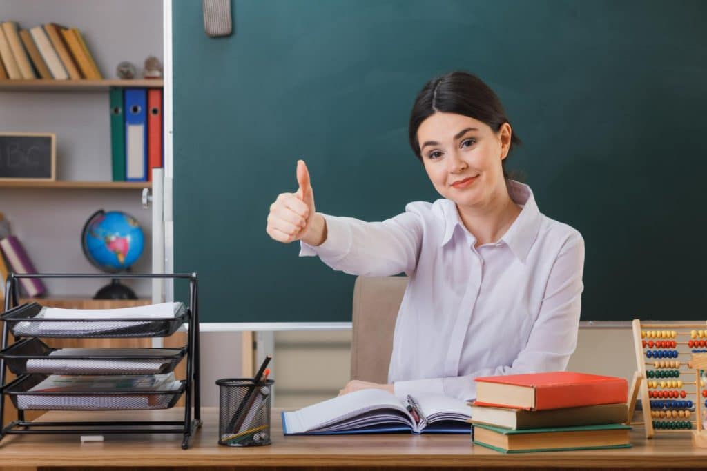 Mulher sorrindo em uma sala de aula, dando um sinal de positivo enquanto está em frente ao quadro negro.