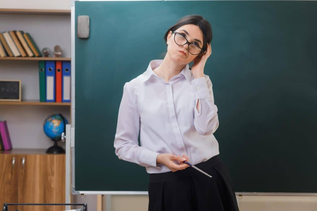 Professora de óculos está pensativa em frente ao quadro negro em sala de aula