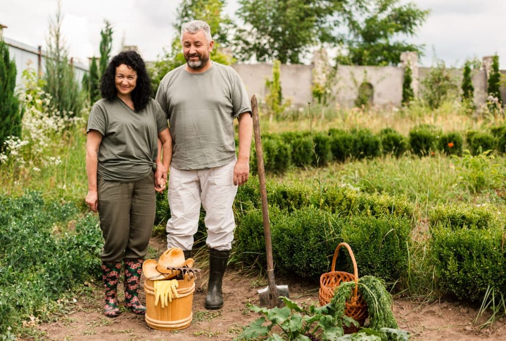 Casal sorridente em uma horta orgânica, com ferramentas de jardinagem e cestos de colheita.