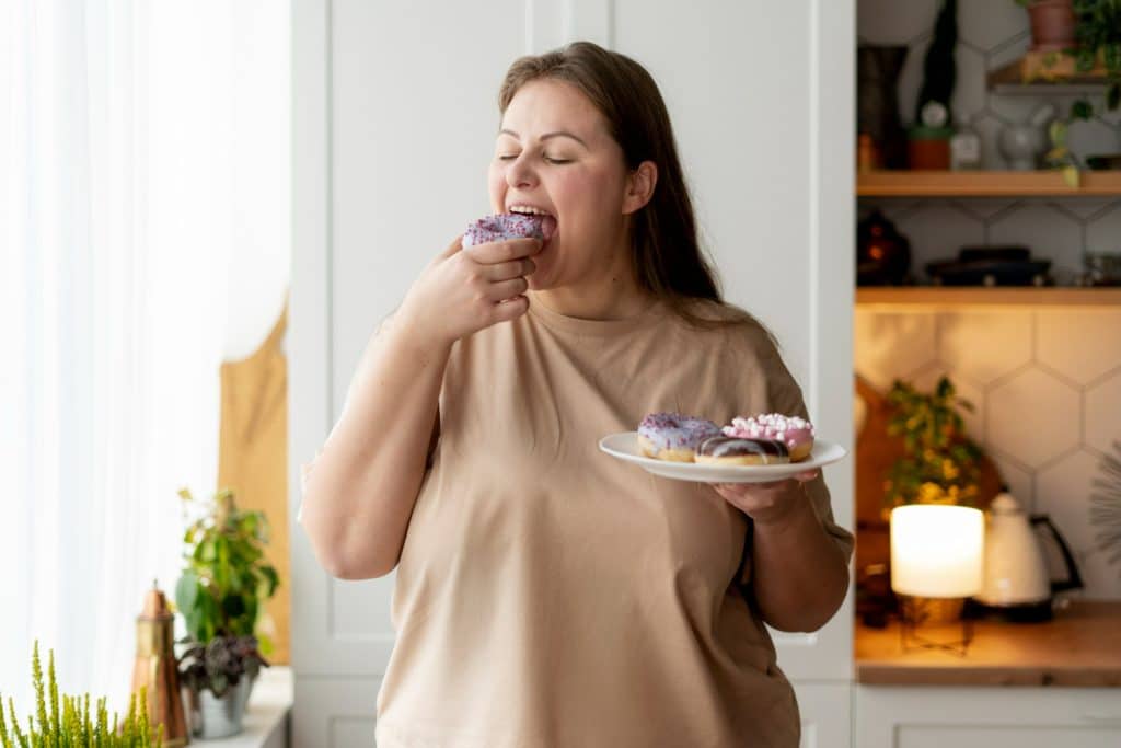 Mulher feliz comendo donut roxo com confeitos, segurando prato com mais donuts na cozinha.