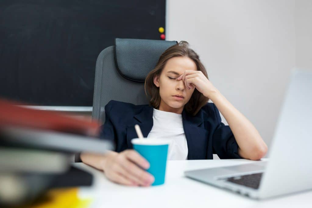 Mulher cansada sentada à mesa de escritório, segurando copo azul, laptop aberto.