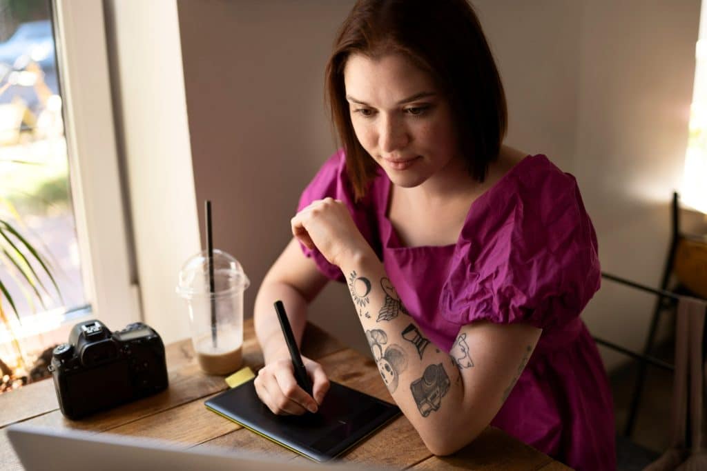 Pessoa canhota desenhando com tablet em ambiente de café, com tatuagens visíveis no braço.