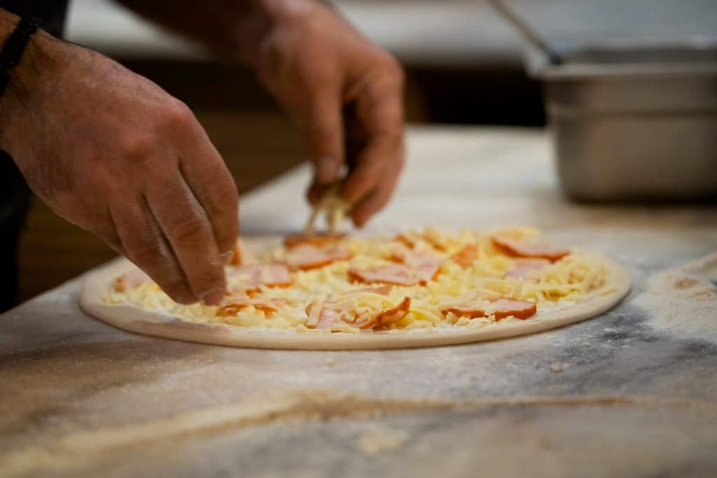 Mãos adicionando ingredientes frescos na massa de pizza artesanal em bancada enfarinhada.