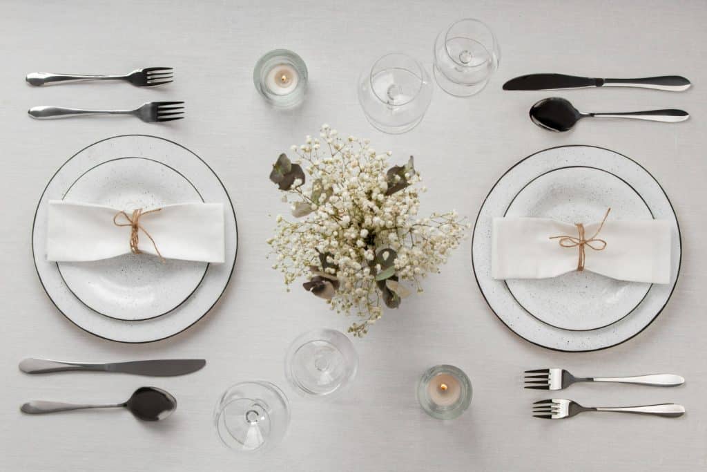Mesa posta para dois com pratos brancos, taças, talheres, velas e arranjo de flores.