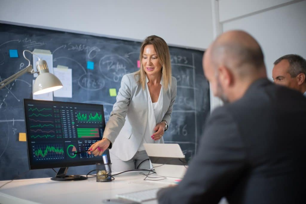 Mulher aponta gráficos em monitor durante reunião empresarial com equipe analisando informações.