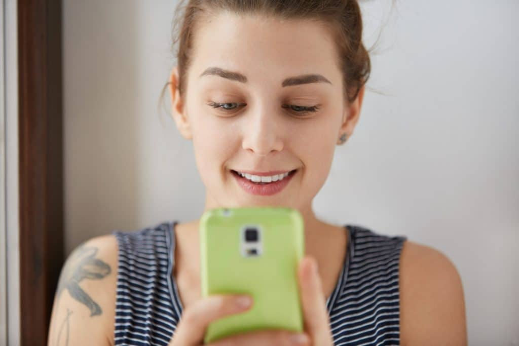 Mulher jovem de cabelo preso sorrindo enquanto olha para celular com capa verde.