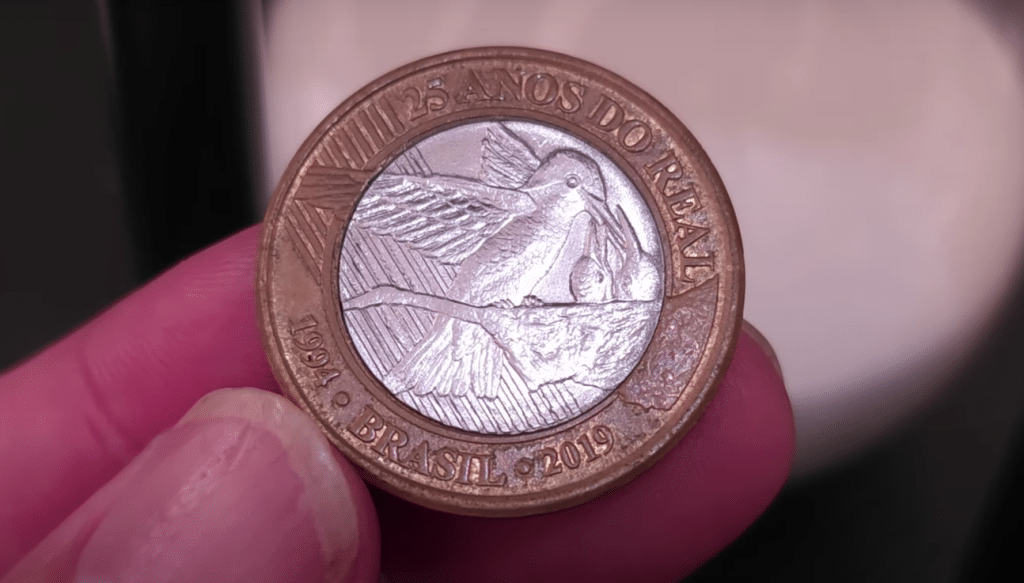 Close-up de uma moeda comemorativa de 1 real, mostrando a efígie do beija-flor no centro e as inscrições "25 ANOS DO REAL", "1994" e "2019" na borda.