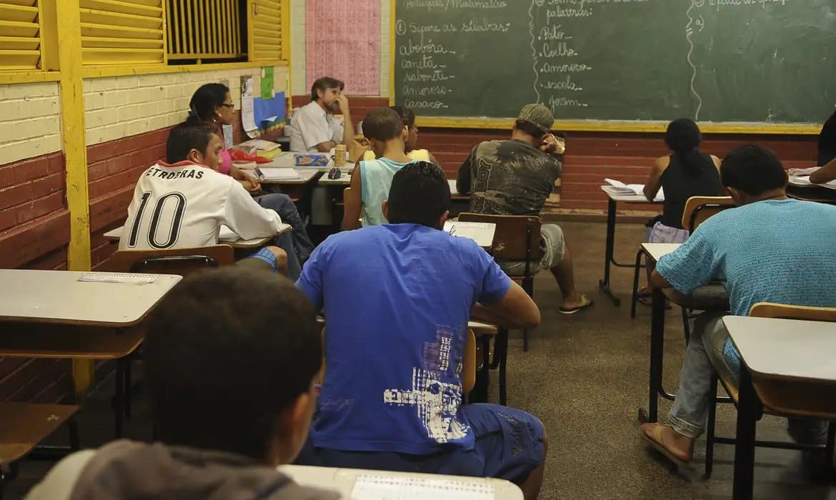 Estudantes adultos assistem aula em sala da EJA durante o período noturno.