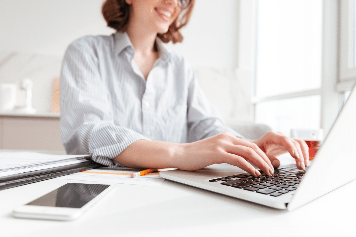 Mulher digitando em notebook em ambiente claro com celular na mesa
