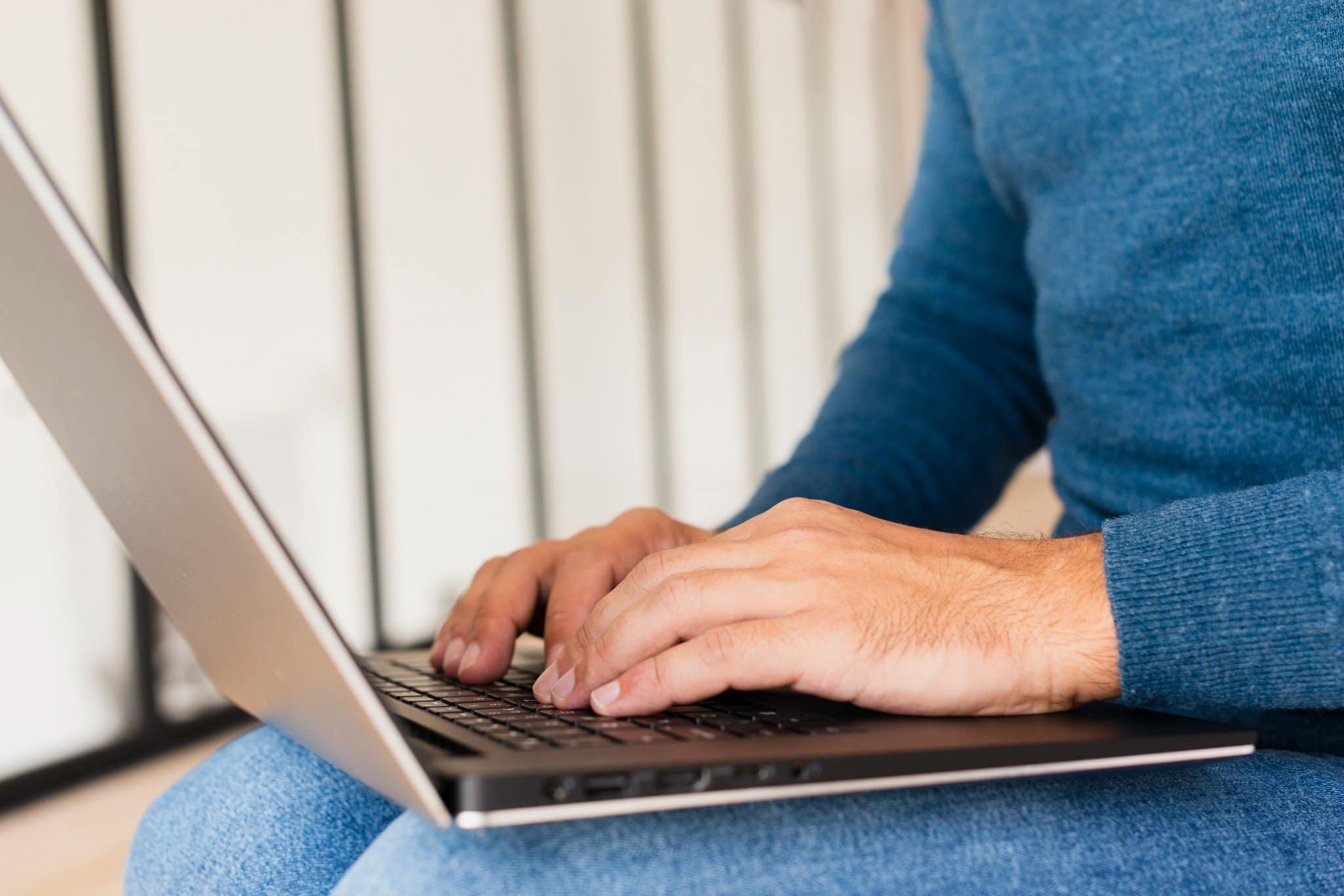 Homem digitando em notebook no colo, usando roupa azul.