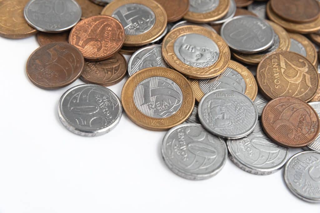 Conjunto de moedas brasileiras de diferentes valores (1 real, 50 centavos, 25 centavos, 10 centavos e 5 centavos) espalhadas sobre um fundo branco, sugerindo a busca por itens valiosos.