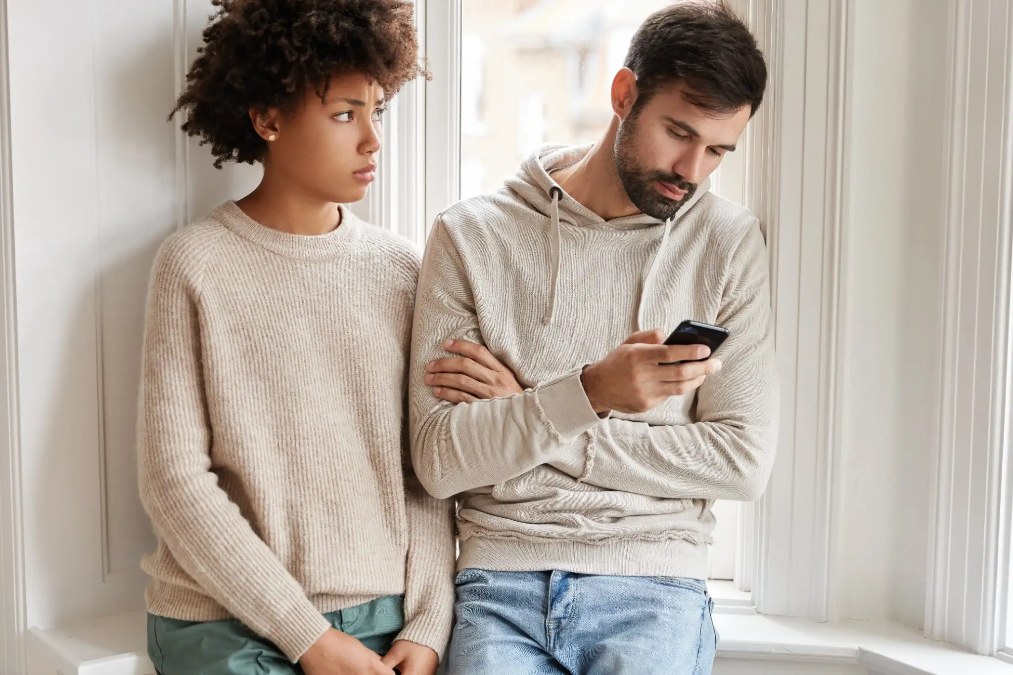 Mulher triste em suéter observando seu namorado distraído no celular, refletindo sinais de indiferença no relacionamento.