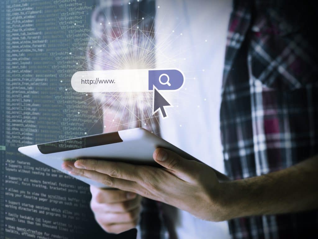 Pessoa segurando um tablet com uma barra de pesquisa e um cursor, simbolizando a navegação na web e a escolha de domínio.