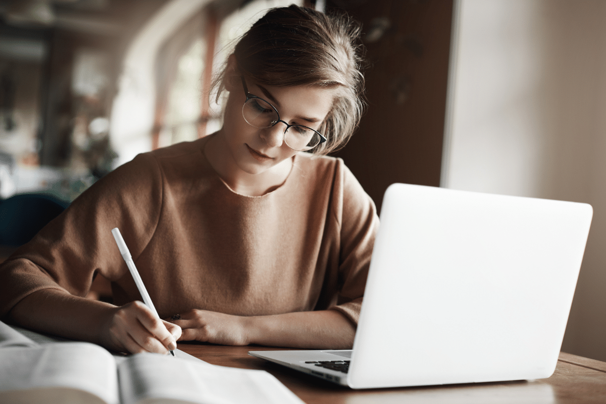Jovem estudando com notebook e livro aberto em mesa de madeira.