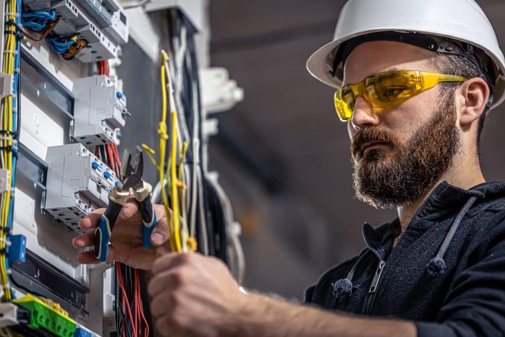 Um eletricista usa óculos de proteção e um alicate para conectar cabos em um quadro de distribuição elétrica.