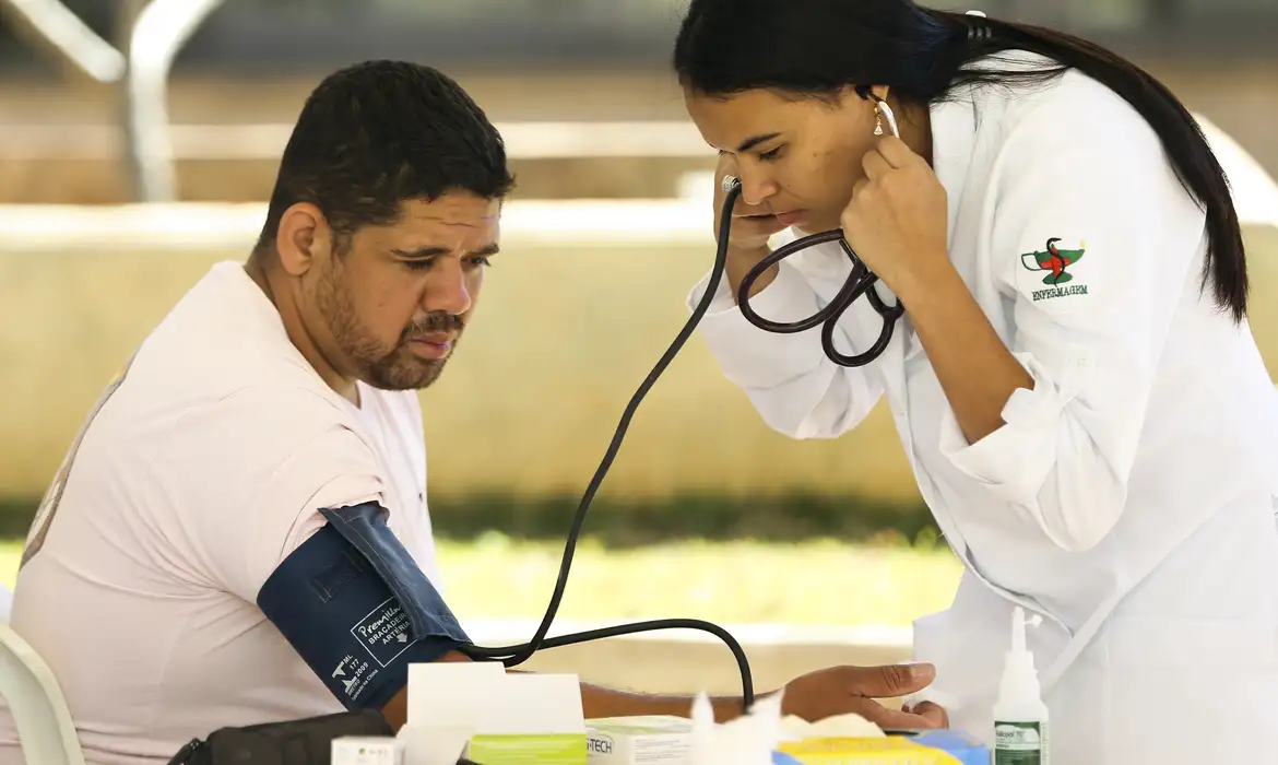 Homem sendo examinado por uma profissional de saúde, medindo a pressão arterial, representando a inspeção médica oficial de concursos públicos.