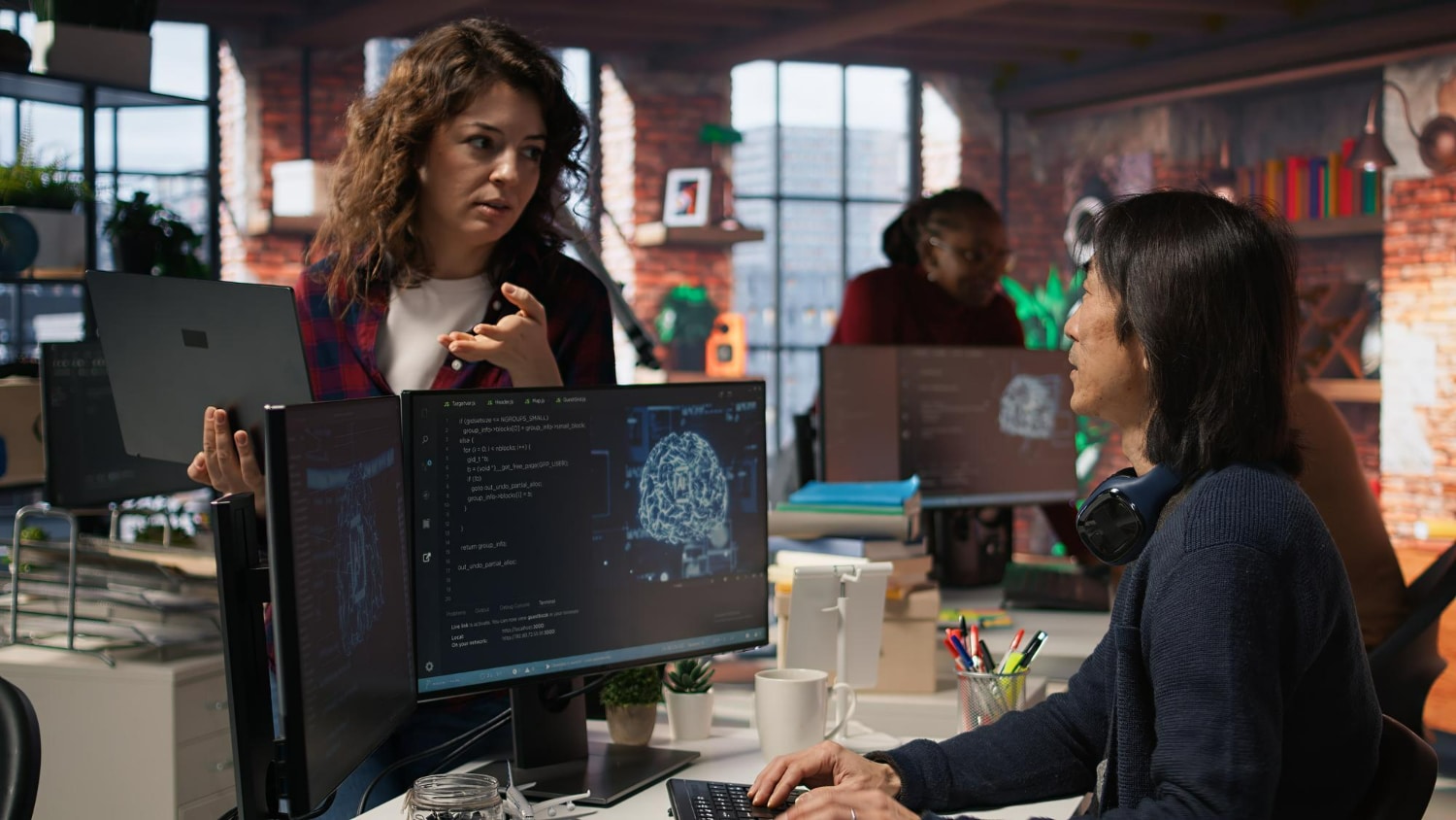 Duas pessoas em escritório discutindo código em computadores com imagens digitais de cérebro na tela.