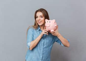 Jovem mulher com camisa azul sorridente segurando cofrinho rosa em formato de porquinho.