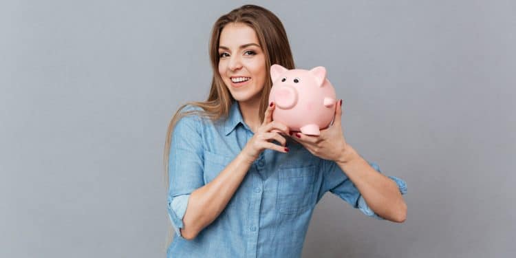Jovem mulher com camisa azul sorridente segurando cofrinho rosa em formato de porquinho.