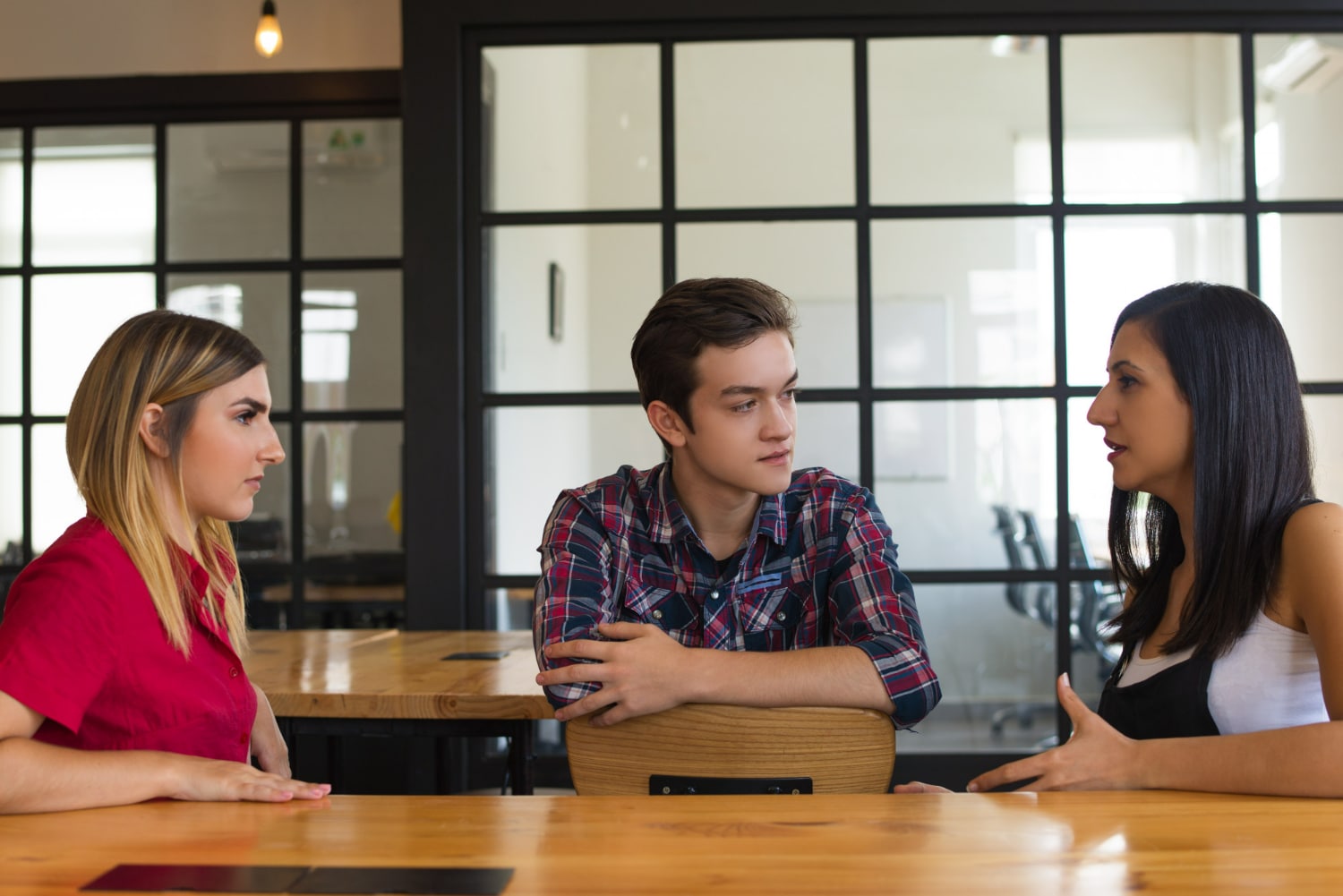 Três jovens conversando de forma séria e atenta em sala moderna com mesa de madeira.