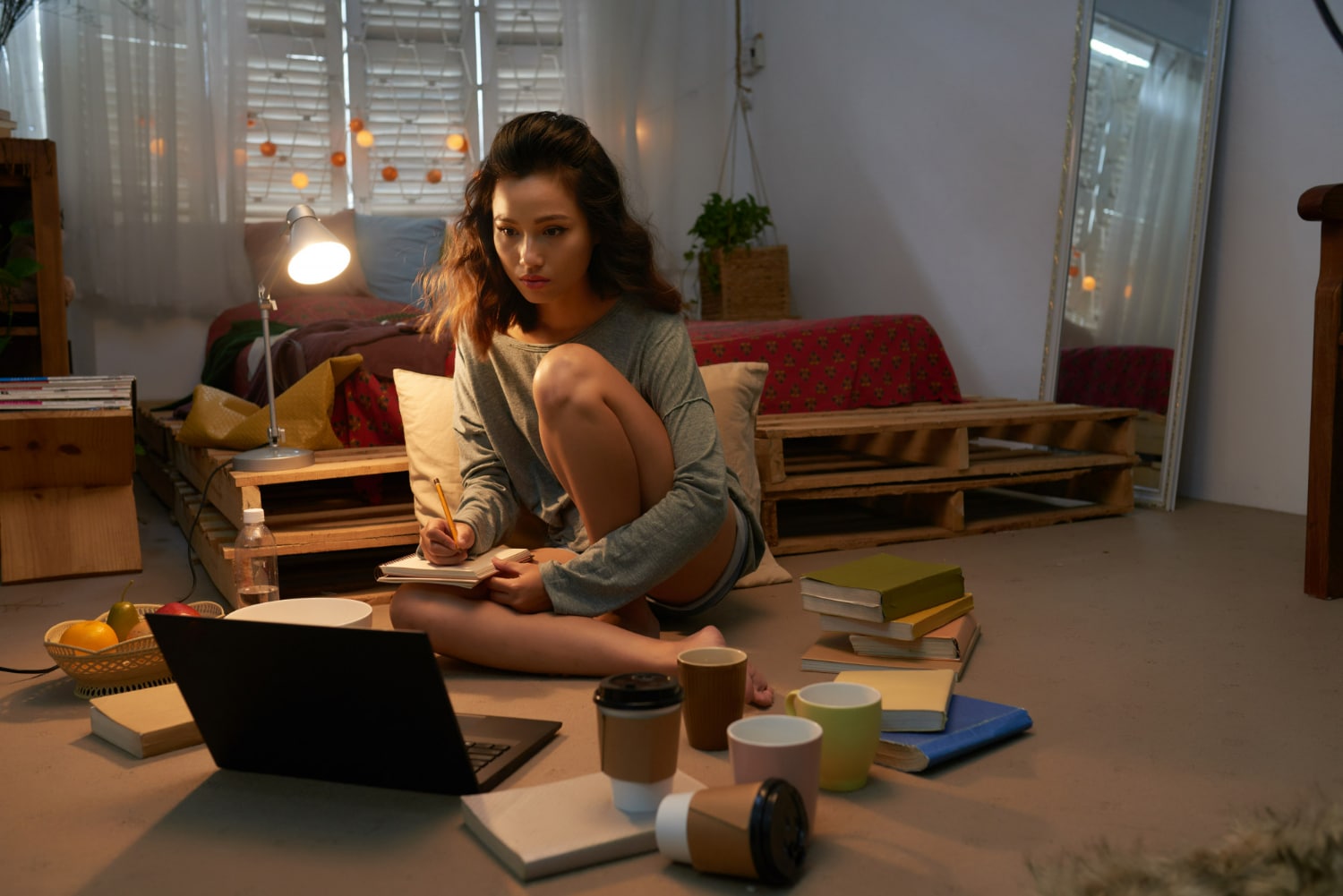 Mulher sentada no chão estudando com laptop, cadernos, livros e várias xícaras em ambiente aconchegante.