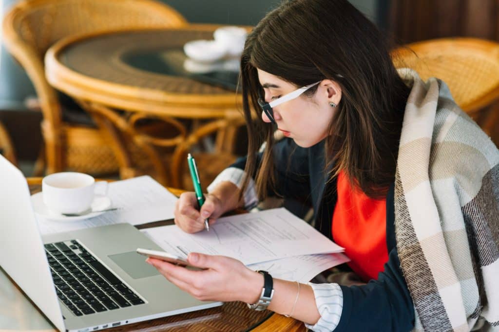Mulher usando óculos escrevendo em documentos enquanto segura celular, com laptop e xícara na mesa.