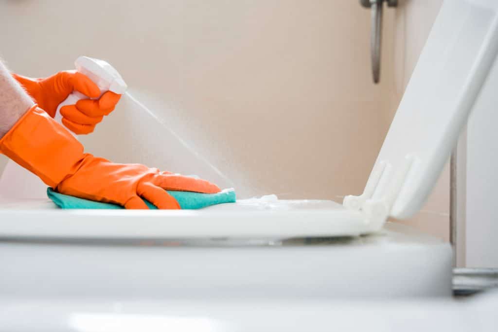 Pessoa com luvas laranja aplicando spray limpador e usando pano azul para higienizar vaso sanitário branco.