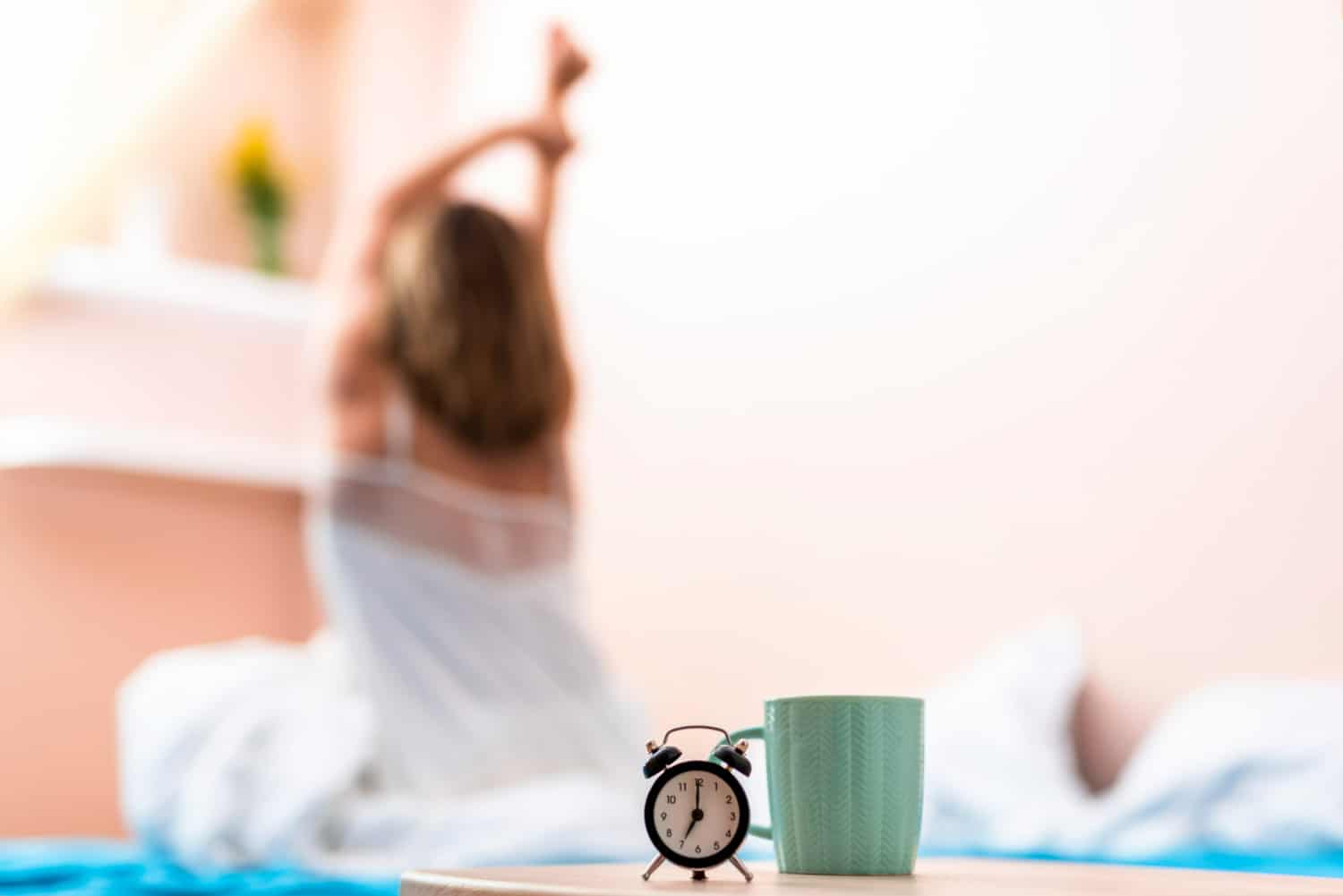 Despertador preto e caneca verde sobre mesa com mulher alongando no fundo desfocado.