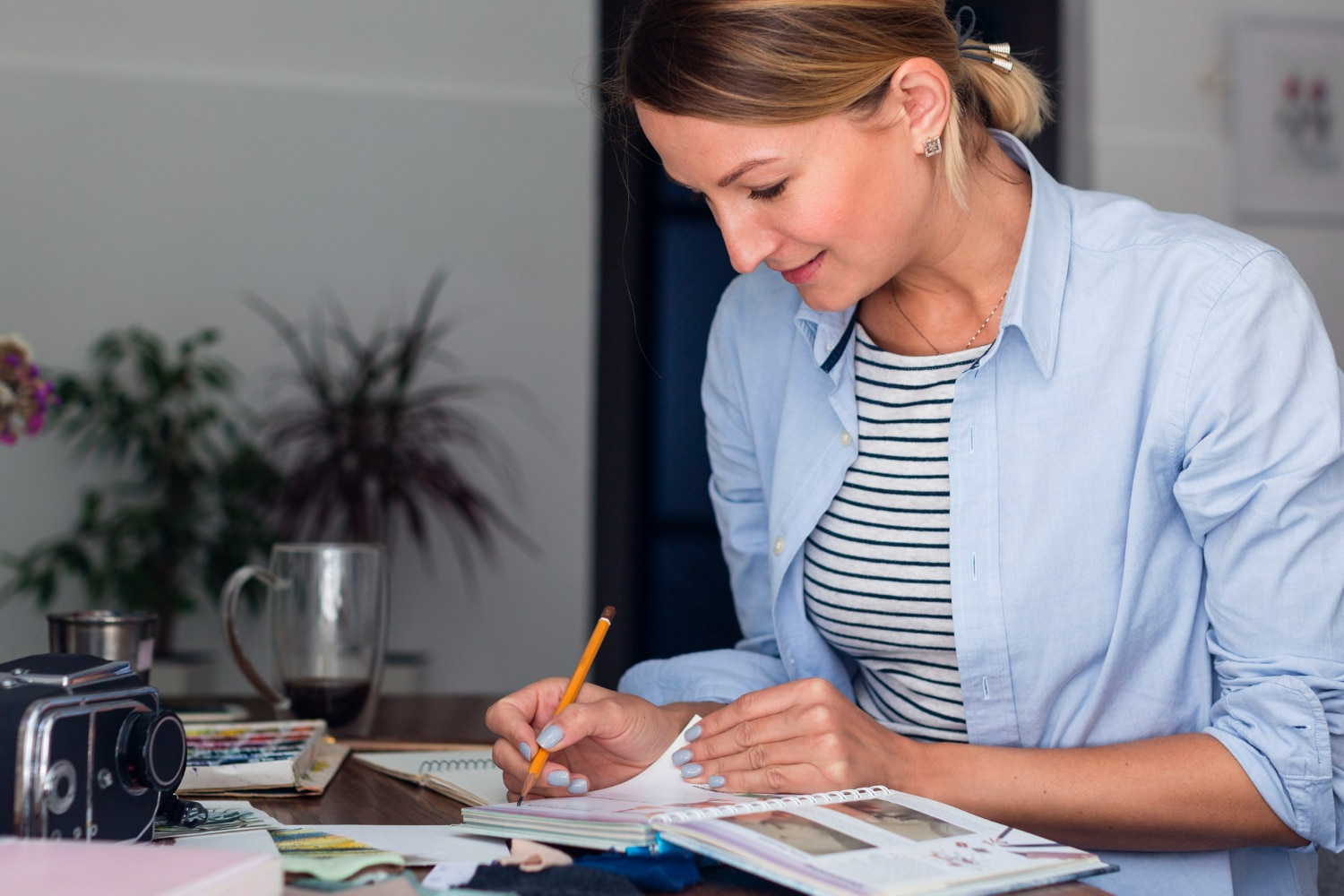 Mulher jovem anotando ideias em diário, com câmera e materiais de arte na mesa.
