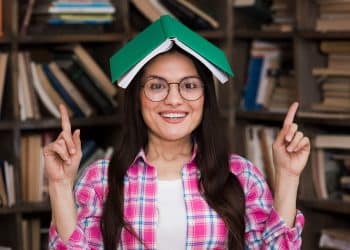 Mulher jovem usando óculos e camisa xadrez rosa com livro verde aberto sobre a cabeça em biblioteca.