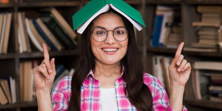 Mulher jovem usando óculos e camisa xadrez rosa com livro verde aberto sobre a cabeça em biblioteca.